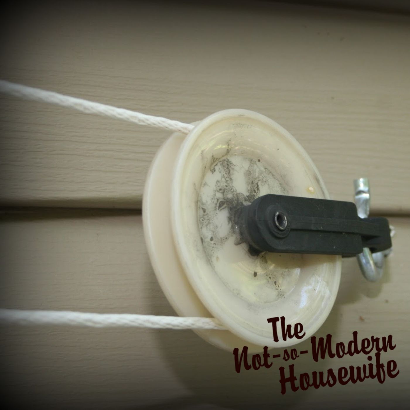 A close-up of a pulley clothesline installation mounted on the side of a house with a nylon clothesline rope threaded through the pulley. The pulley is secured with a steel screw hook, showing a practical example of an outdoor clothesline system for energy-efficient laundry solutions. The image highlights the simplicity and functionality of a DIY clothesline project, a staple in Christian homemaking and sustainable living. The branding of The Not-So-Modern Housewife is visible, reinforcing a commitment to faith-based simple living and homesteading practices.