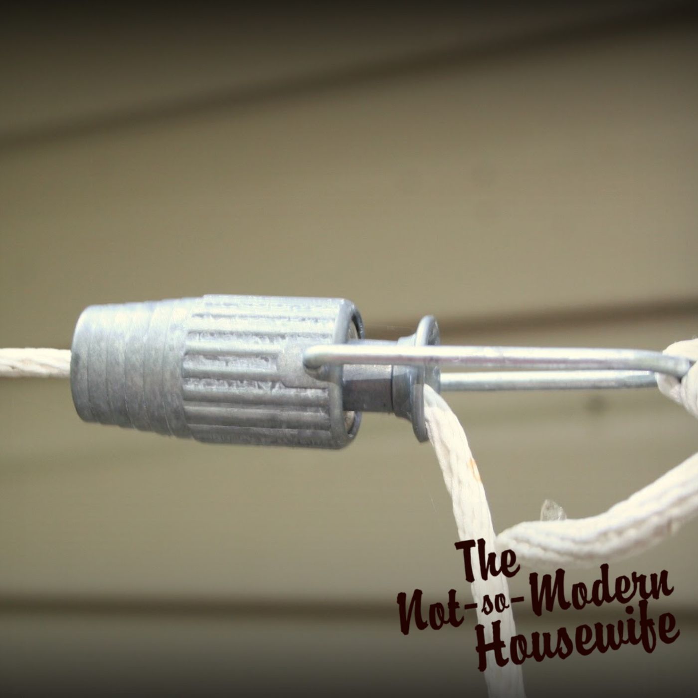 A close-up of a clothesline tightener with a ball-bearing mechanism, securely holding a nylon clothesline rope in place. This clothesline tension adjuster allows for easy tightening as the rope stretches over time, ensuring an efficient outdoor clothesline system. Installed near the house, this DIY laundry solution supports energy-efficient and eco-friendly homemaking by reducing electricity use. The branding of The Not-So-Modern Housewife reinforces a commitment to faith-based simple living and sustainable homesteading practices.