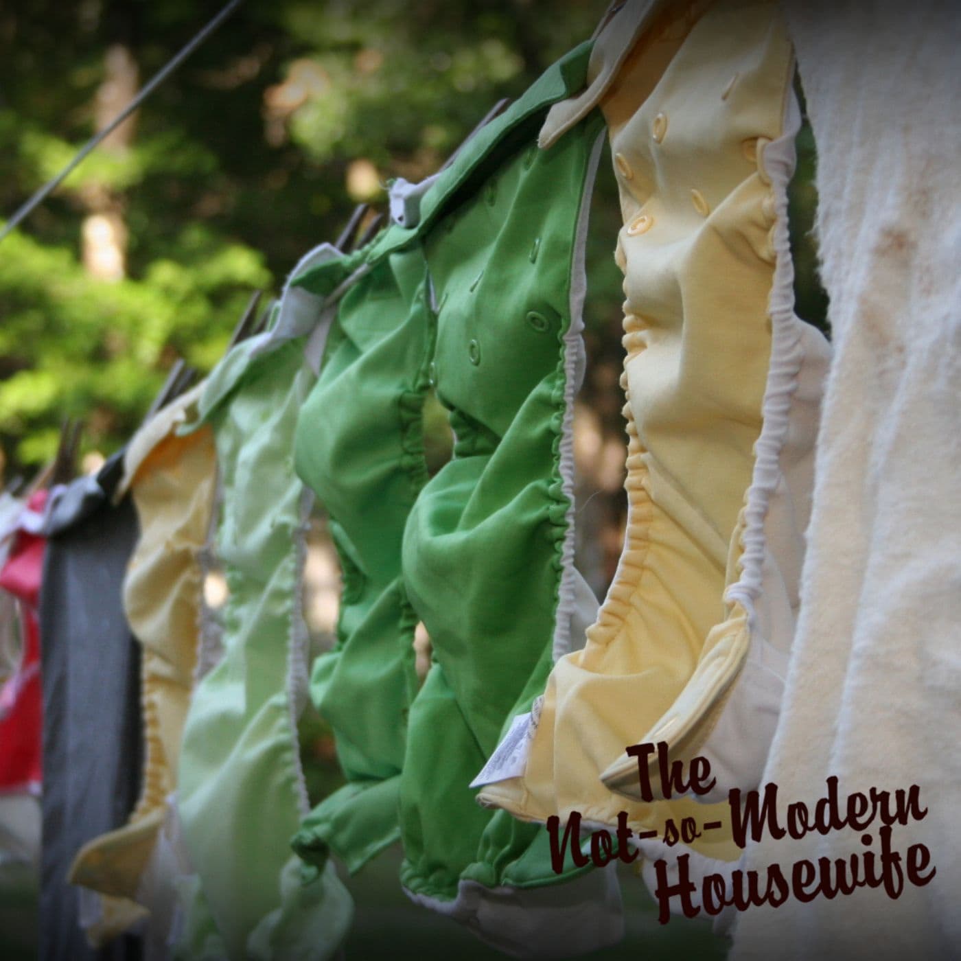 A row of cloth diapers in shades of green, yellow, and red drying on a pulley clothesline outdoors. This eco-friendly laundry solution helps reduce electricity use while extending the lifespan of reusable diapers. The DIY clothesline setup is part of a sustainable homemaking routine, promoting energy-efficient and natural fabric care. The image showcases the simplicity and practicality of faith-based simple living and homesteading practices, as emphasized by The Not-So-Modern Housewife branding.