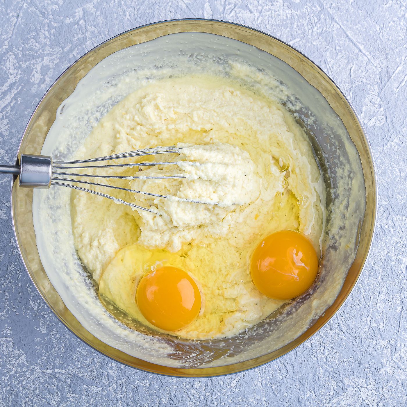 A stainless steel mixing bowl with cake batter, two cracked eggs, and a whisk, showcasing the process of making box cake mix with pudding. The smooth, rich batter highlights how instant pudding mix enhances moisture and texture, creating a soft and flavorful cake. Perfect for a quick, homemade dessert that tastes bakery-quality with minimal effort.