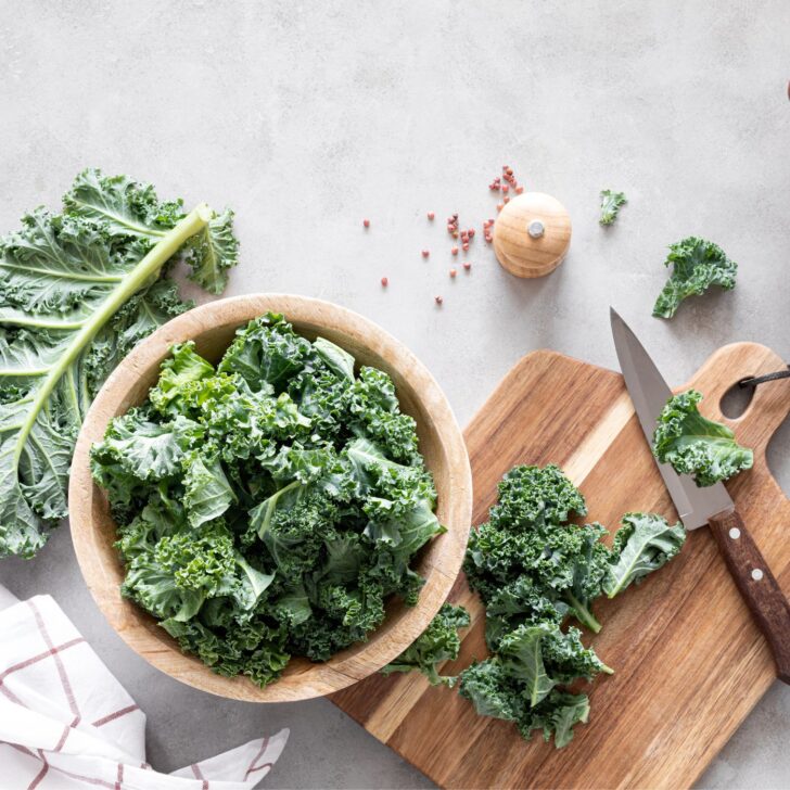 Hearty Potato and Kale Frittata Recipe: A Delicious and Nutritious Dish for Any Meal | The Not so Modern Housewife Fresh kale leaves in a wooden bowl, surrounded by a cutting board, a knife, and a neatly arranged kitchen towel, set on a light gray countertop – a simple and inviting setup for meal prep.