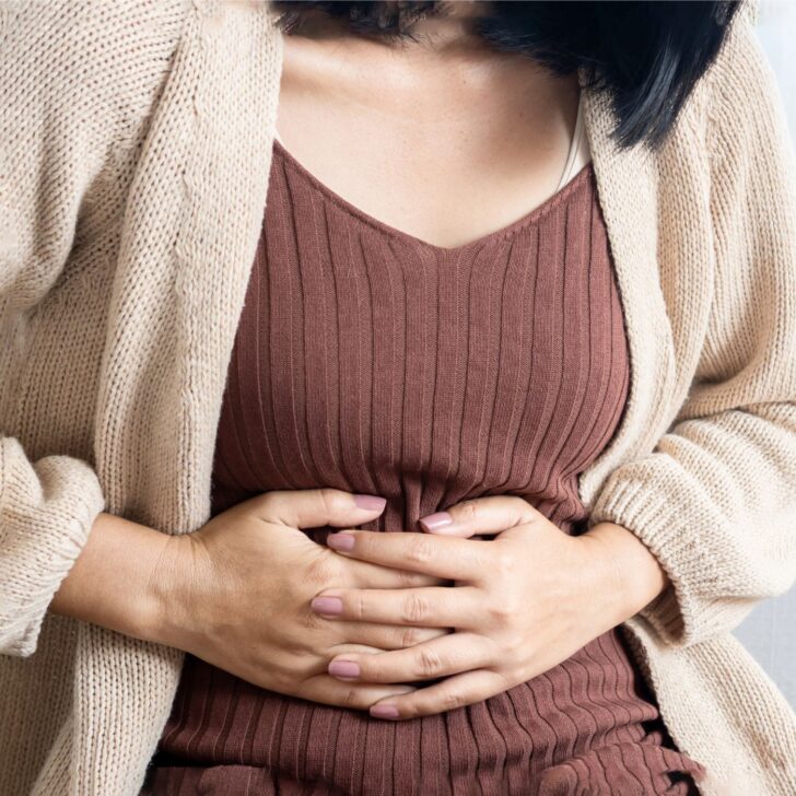 Understanding and Managing Estrogen Dominance Naturally | The Not so Modern Housewife woman with a brown top and tan cardigan holding her stomach - signs of estrogen dominance