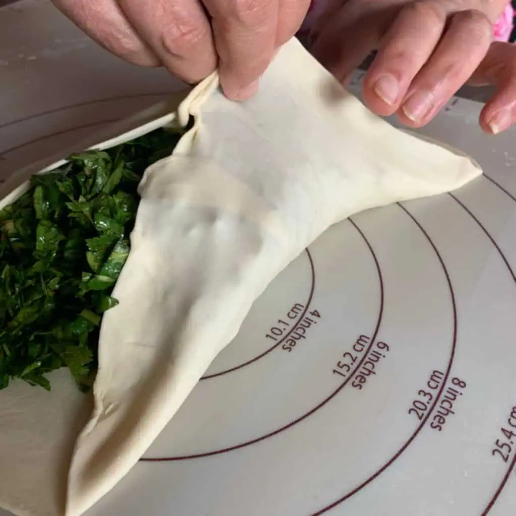 Two cups of seasoned mixed greens mounded in the center of a rolled-out flatbread dough circle, ready to be folded and sealed.