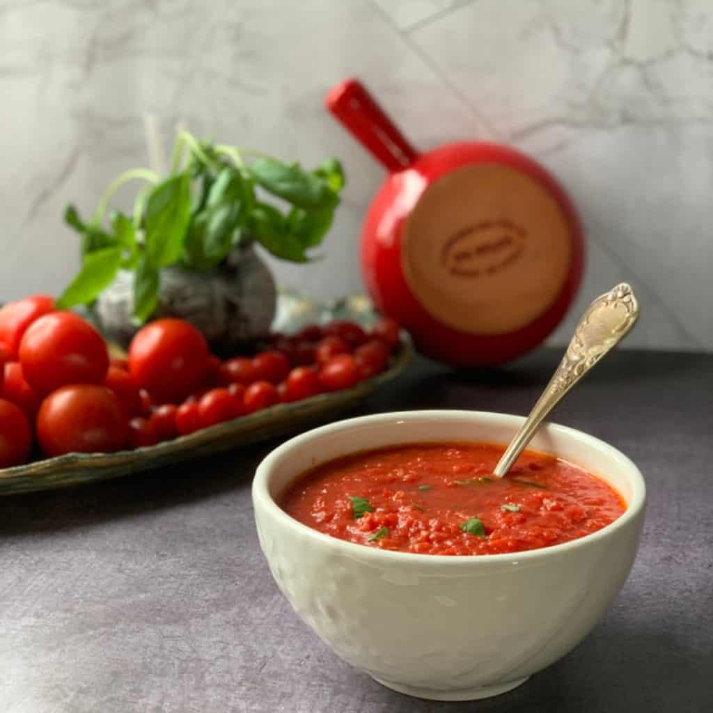 Sugo in a white bowl and tomatoes and basil plant in a background.