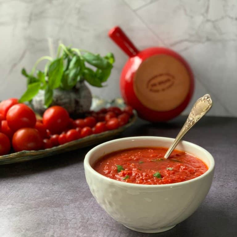 Sugo in a white bowl and tomatoes and basil plant in a background.