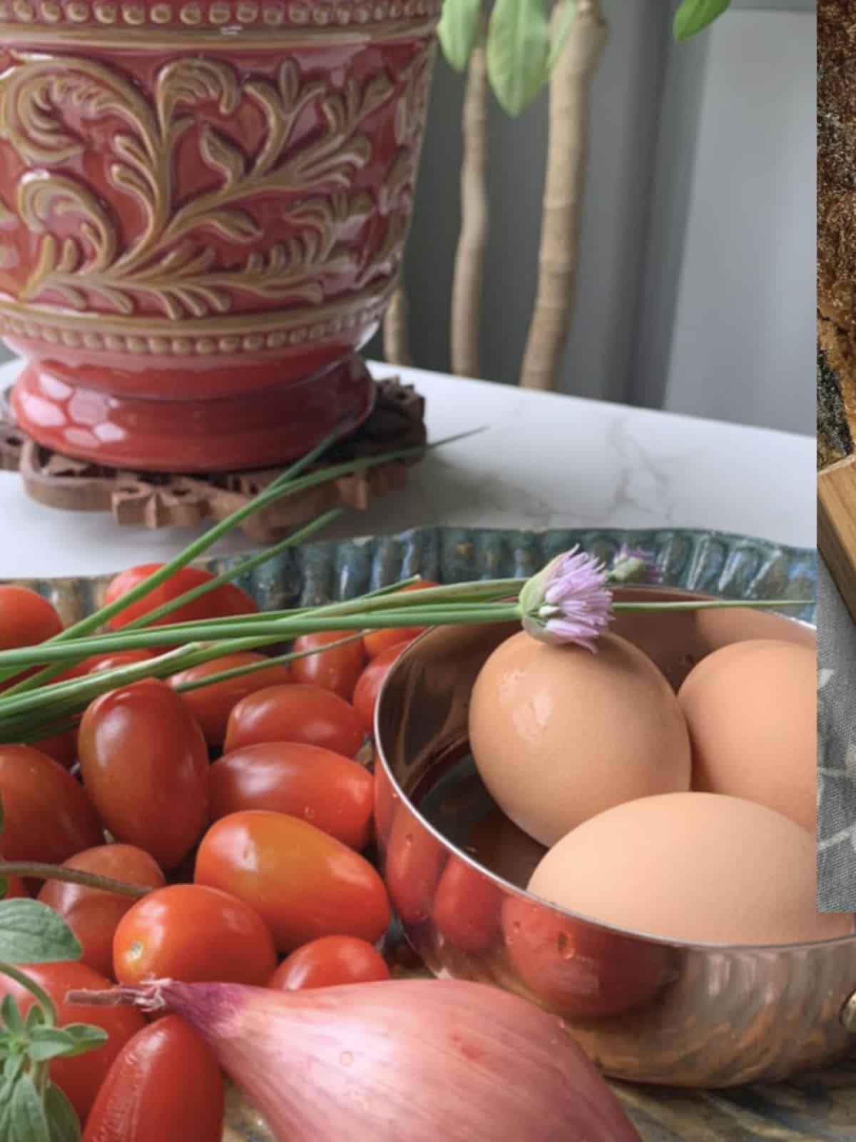 Grape tomatoes, eggs, chives, and shallots in a foreground and a red planter in the background.