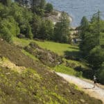 Hiker walking along a trail with lush green hills and lake in the background.