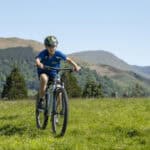 Child riding a mountain bike on grassy field with hills in background.