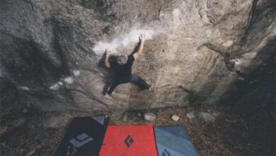 Climber scaling a steep outdoor rock face with crash pads below.