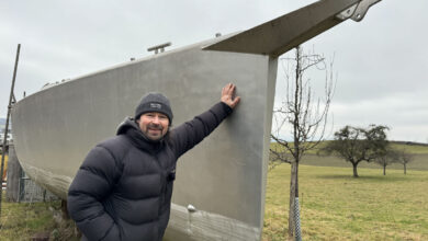 Boat hull under repair outdoors in a grassy field with trees, man inspecting boat.