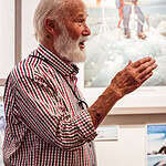 Older man with white hair and beard explaining outdoor adventure photography at a gallery.
