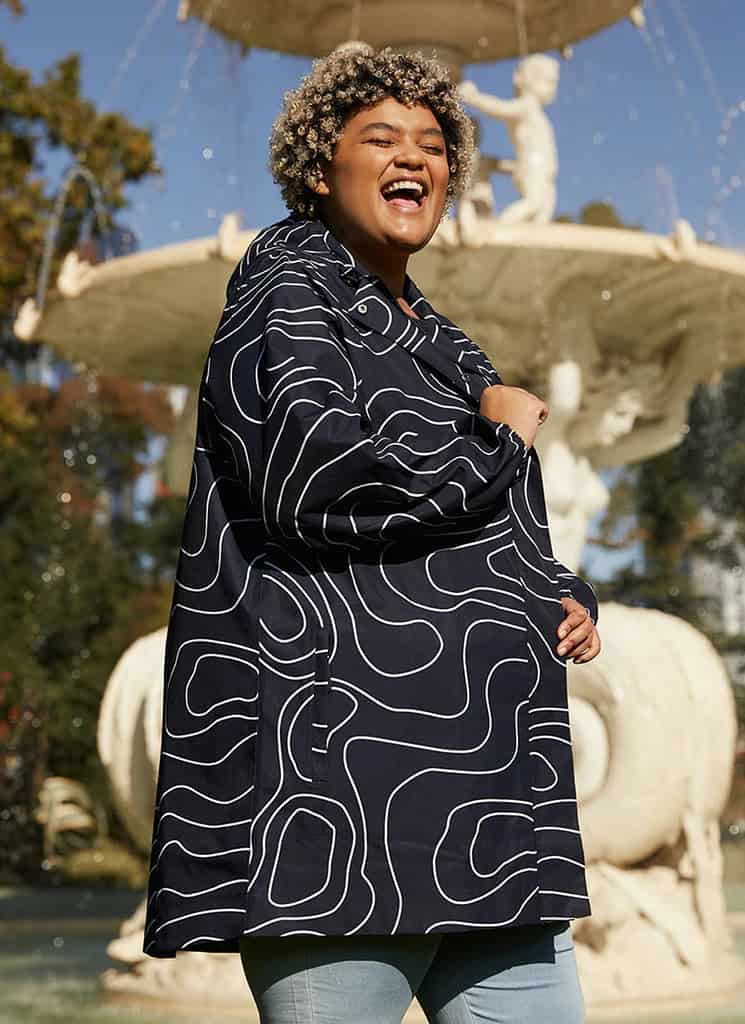 Bright woman wearing stylish waterproof jacket outdoors near fountain, perfect for outdoor adventure clothing.