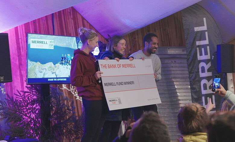 People celebrating outdoor adventure award win on stage at Merrell event in cosy wooden venue, smiling with large cheque.