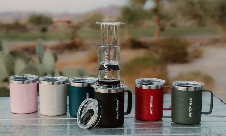 Reusable coffee cups and a glass coffee press on a wooden table outdoors.