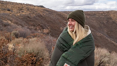 Woman in warm sleeping bag outdoors in rugged landscape.