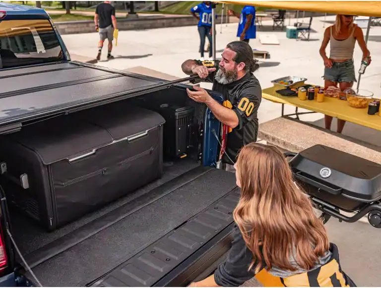 a man and a woman unpacking items from the back of a blue pickup truck. The truck bed contains a large, black rectangular storage bag and a smaller, black rectangular box. This image is showcasing the BakFlip MX4 Hard Folding Tonneau Cover