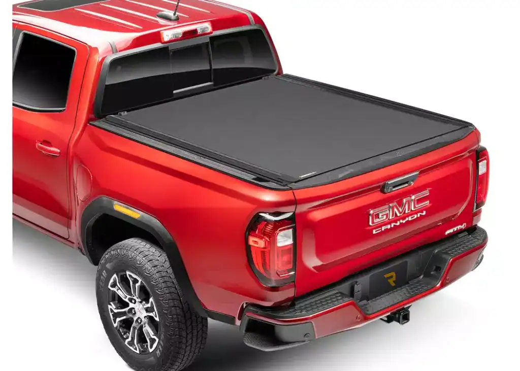 A high-angle studio shot showcases the rear three-quarter view of a red GMC Canyon pickup truck isolated on a white background. The truck bed is completely sealed with a low-profile, matte black retractable tonneau cover that sits flush with the bed rails. The vehicle features black trim along the rear bumper and wheel wells, large silver and black multi-spoke wheels, and distinct red taillights. The chrome "GMC CANYON" emblem is prominently displayed on the tailgate, and a dark license plate with a yellow geometric "R" logo sits above the hitch receiver.