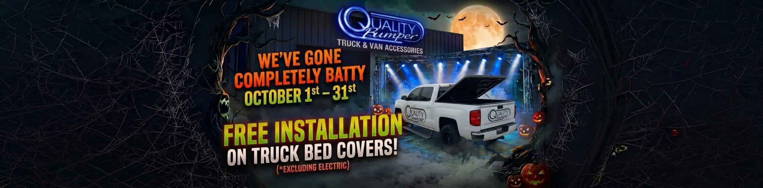 A Halloween-themed promotional graphic for Quality Bumper features a white pickup truck parked in front of a store under a full moon, lit by blue spotlights and surrounded by fog, jack-o'-lanterns, bats, and spiderwebs. The store building displays a glowing blue "Quality Bumper TRUCK & VAN ACCESSORIES" logo sign. The pickup truck features Quality Bumper door and tailgate decals and has an open tonneau cover on its bed