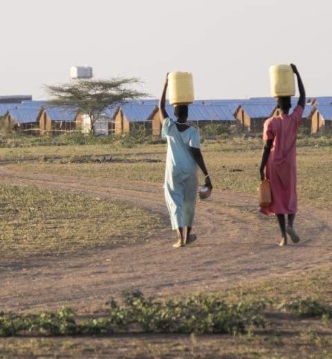 Two women carrying yellow water containers on their heads along a dirt path