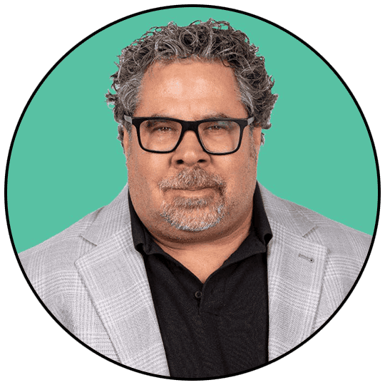 A professional man in a grey blazer and black shirt, wearing glasses, against a teal background.