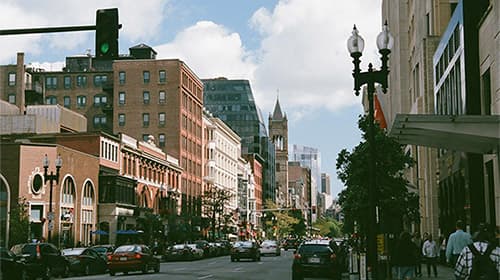 A street in an urban area featuring cars and buildings under a cloudy sky.