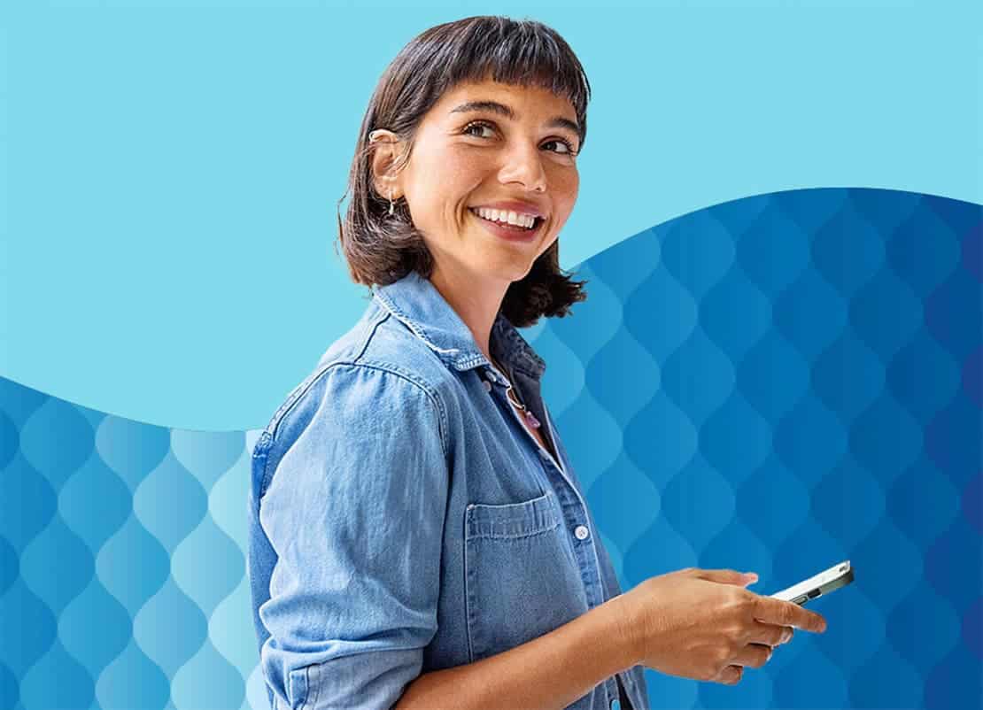 A woman smiling while holding a phone, wearing a blue shirt against a bright sky.