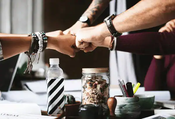 Team members' hands join in a fist bump over a desk with office supplies.