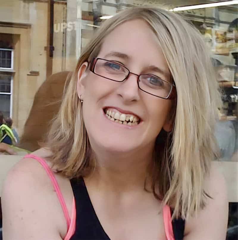 Fran whitaker of the happy journals, with blonde hair, glasses, and a black tank top with pink straps, smiles while sitting outside a café. The background shows a window with reflections and a person in a yellow vest.