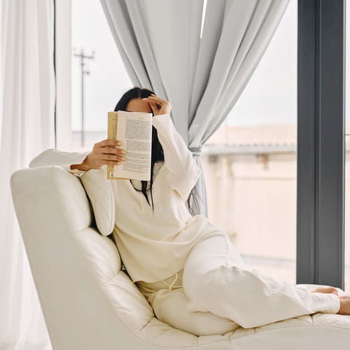 A woman with long brown hair, dressed in a cream tracksuit, reading a book on morning journaling