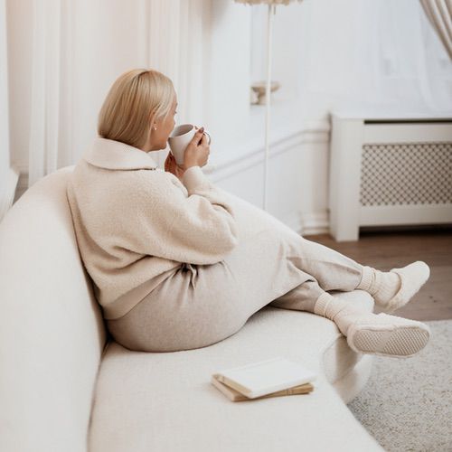 A woman with blonde hair sits on a white sofa, wearing cozy cream-colored loungewear and socks, holding a mug. An open book about journaling for beginners lies beside her. The room has soft lighting and a calm, minimalist decor.