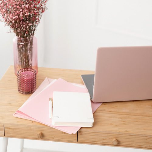 A light wooden desk with a pink laptop, white notebook, pink folders, a pen, a pink candle, and a vase of small pink flowers—perfect inspiration if you’re learning how to make a guided journal in a soft, feminine workspace.