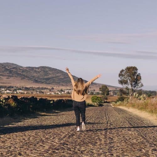 A person with long hair walks down a rural dirt road, arms raised in a gesture of confidence boosters, surrounded by fields and distant hills under a clear sky.