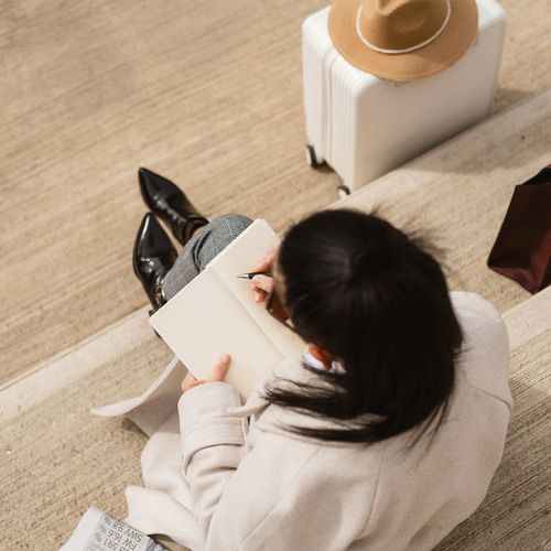 A person with long dark hair sits on outdoor steps, writing in a notebook—perhaps capturing tips on how to travel journal. Nearby are a white suitcase with a tan hat on top and a brown bag. The individual wears a light coat and black boots.