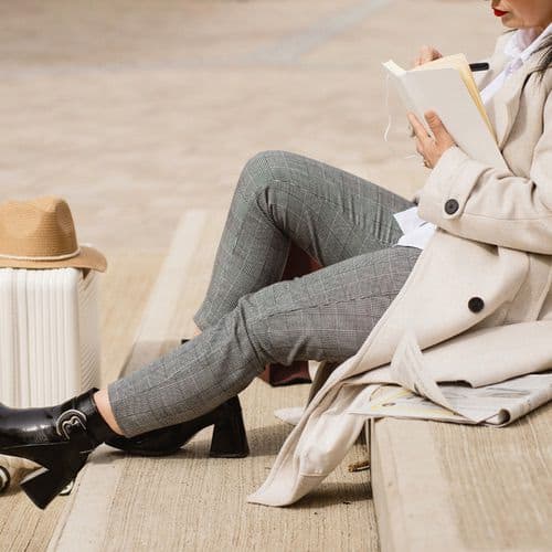 A person in grey plaid pants, a light coat, and black heeled boots sits on outdoor steps, reading about how to travel journal. A tan hat and white suitcase nearby suggest the anticipation of a new adventure.