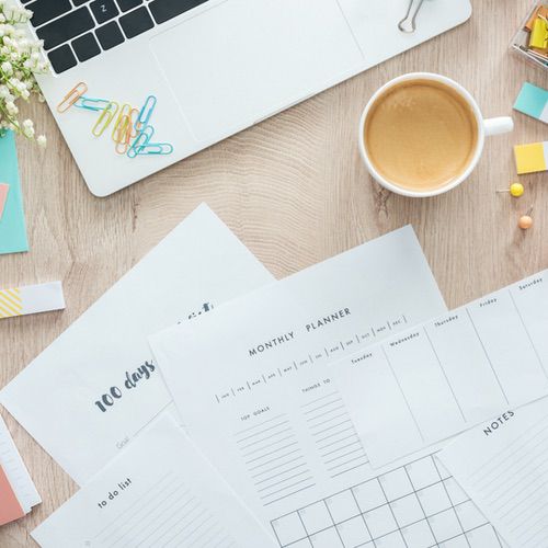 A workspace with a laptop, cup of coffee, paper clips, sticky notes, and several planner sheets—including a monthly planner, to-do list, and notes—showcasing different types of planners for journaling on a wooden desk.