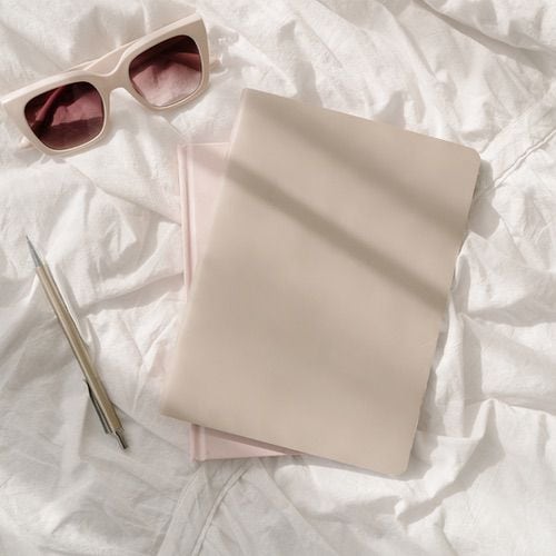 Two closed beige and pink notebooks lie on white wrinkled fabric beside a silver pen and light pink sunglasses, all bathed in soft, natural sunlight—perfect essentials for mindful wellness journaling.