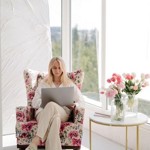 A woman with blonde hair sits on a floral armchair by sunlit windows, using her laptop for wellness journaling. Next to her, a round table holds two vases of pink and white flowers.