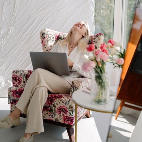 A woman sits in a floral armchair with a laptop on her lap, smiling and leaning back. Next to her is a round table with a vase of pink flowers—perfect for some wellness journaling as sunlight streams in through large windows behind her.
