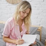 A woman with long blonde hair, wearing pink striped pyjamas, sits on a bed and writes in a blank notebook with a pencil—journalling for improved sleep. She is looking down, focused on her writing, against a white brick wall.