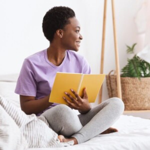 A smiling woman in a purple shirt and grey leggings sits cross-legged on a bed, holding an open yellow notebook—perhaps engaging in nightly journalling to improve sleep—in a bright, cosy room with plants and soft decor.
