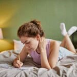 A young woman lies on her stomach on a bed, writing prompts for sleep journalling in a notebook. She wears a light vest and shorts, feet up, with a lamp and green wall visible in the background.