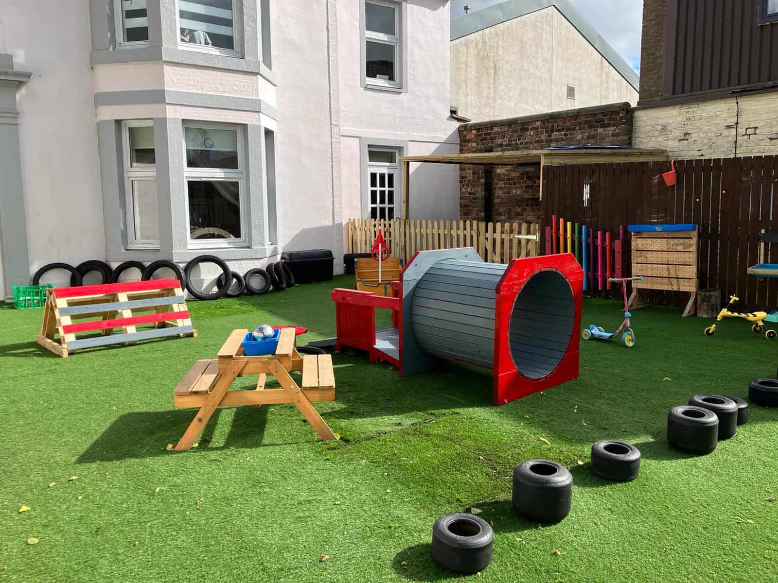 Bright outdoor childcare play area with soft turf, climbing tyres, mini obstacle course, and colourful play structures for children's development and fun.