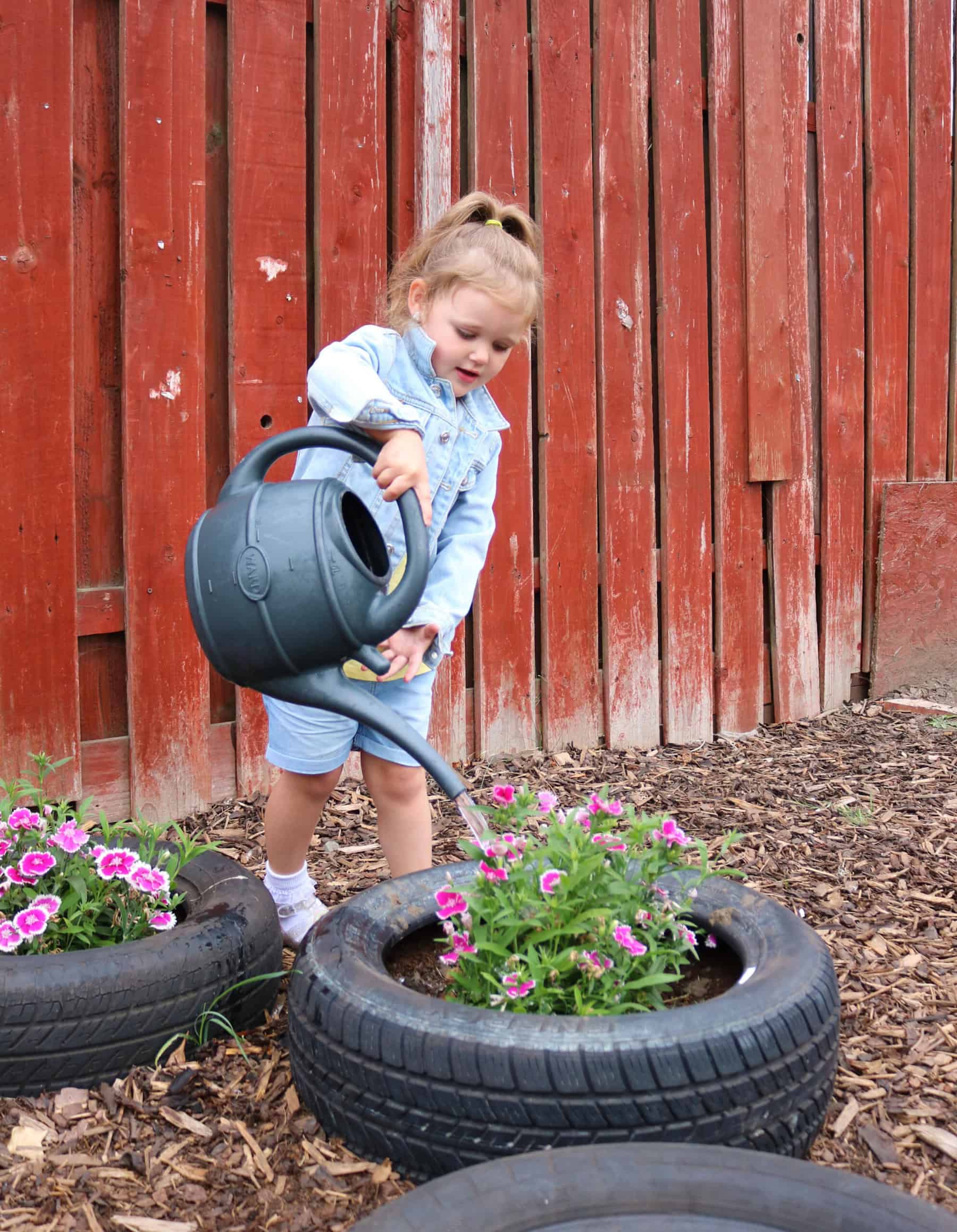 Watering young flowers in a creative outdoor garden with recycled tires at Thrive Childcare. Young girl enjoys outdoor classroom activities, fostering early childhood development and eco-friendly learning.