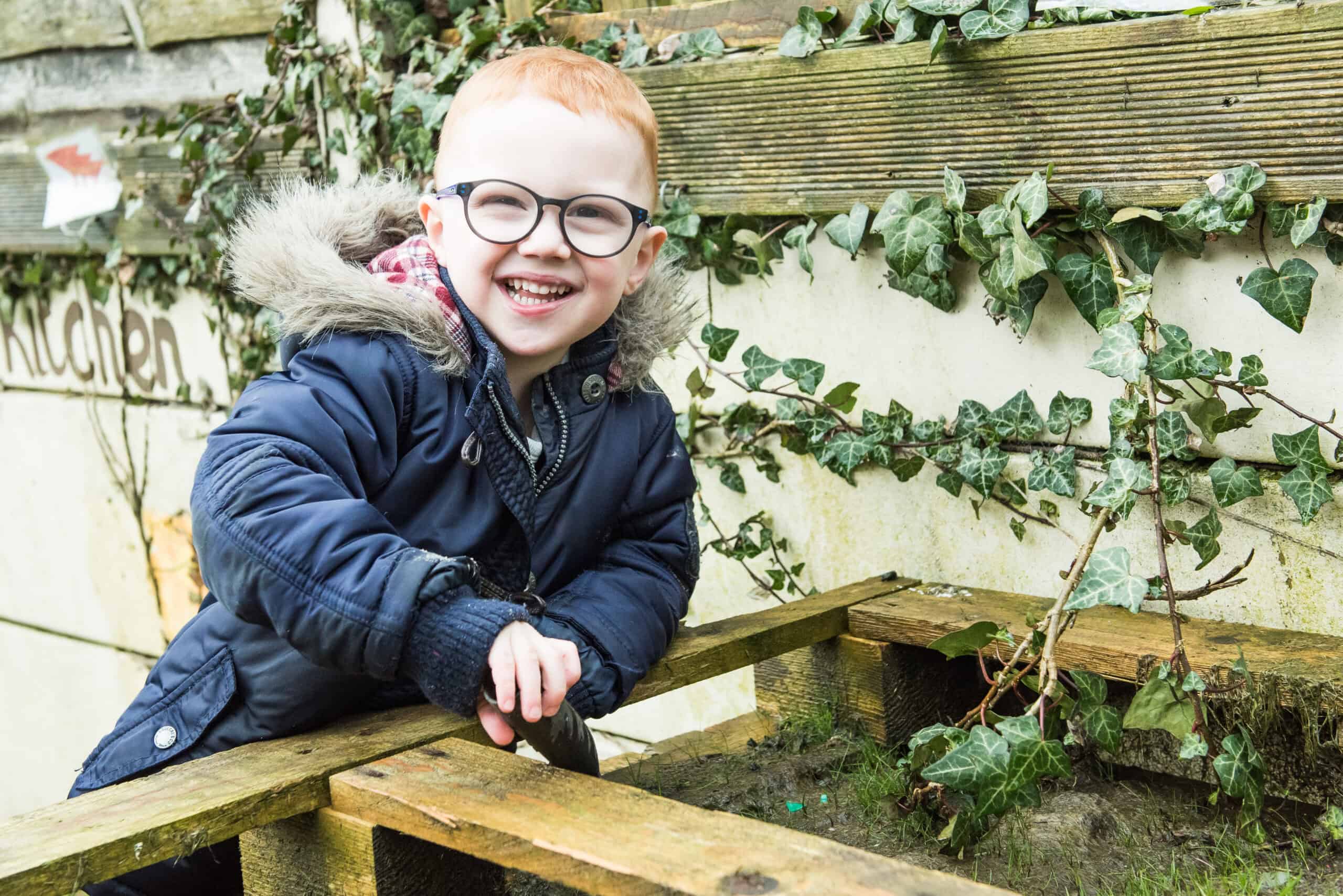 Colourful young boy with glasses gardening outdoors in a childcare setting, engaging in nature-based learning activities at Thrive Childcare.