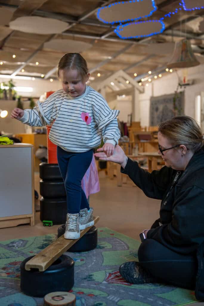 Child balancing on a wooden beam with support from a caregiver in a bright, welcoming nursery environment.