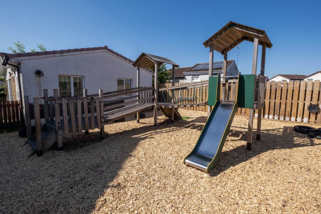 Bright outdoor play area at Thrive Childcare featuring a wooden slide, climbing frame, and safe gravel ground, providing a fun and engaging environment for early childhood development.