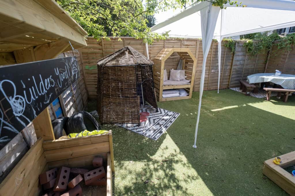 Bright outdoor play area at Thrive Childcare featuring a mini wooden house, wicker play tent, shaded seating, and gardening corner, designed for young children’s outdoor fun and development.