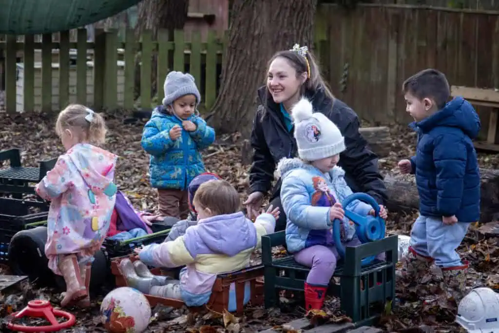Children playing outdoors with caregiver at Thrive Childcare, engaging in imaginative and outdoor activities in a natural play area.