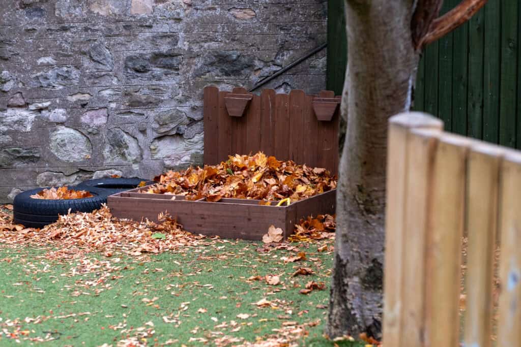 A colourful autumn leaves play area at Thrive Childcare with a wooden leaf collector, tyre and grassy ground, perfect for outdoor learning and child development.