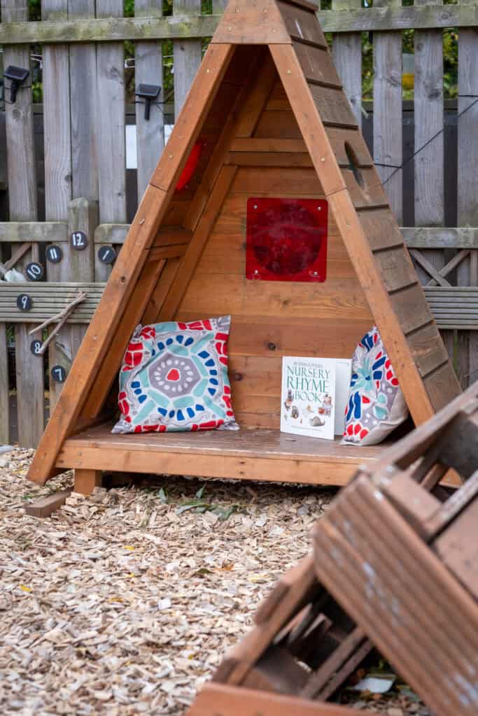 A cosy outdoor reading nook with a wooden teepee, colourful cushions, and a nursery rhyme book, perfect for early childhood development and outdoor play at Thrive Childcare.